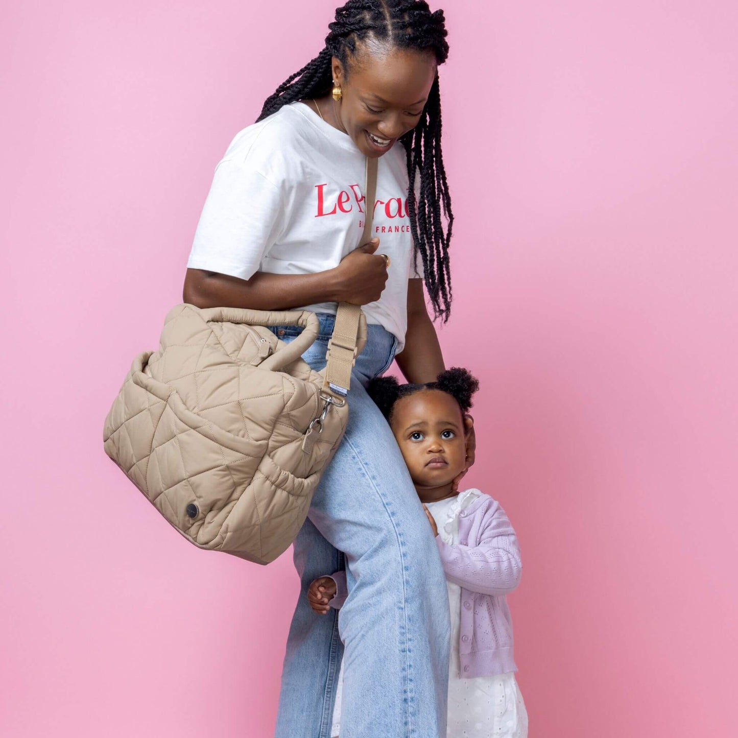 Babymel Willow Convertible Changing Bag in oatmeal, worn crossbody by a smiling mother, with her toddler hugging her leg, highlighting its versatility.