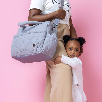 Babymel Willow Convertible Changing Bag in duck egg grey, worn crossbody by a mother while her toddler hugs her leg, showcasing its versatile design.