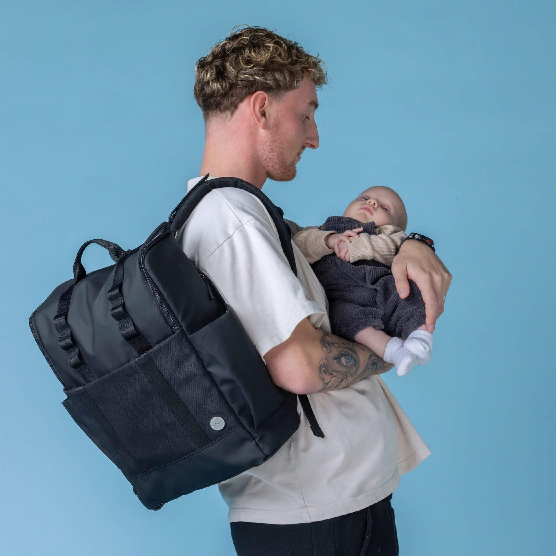 Side view of a parent wearing the Babymel Finn Changing Backpack in Black while cradling a baby, highlighting its ergonomic design and hands-free convenience for everyday parenting.