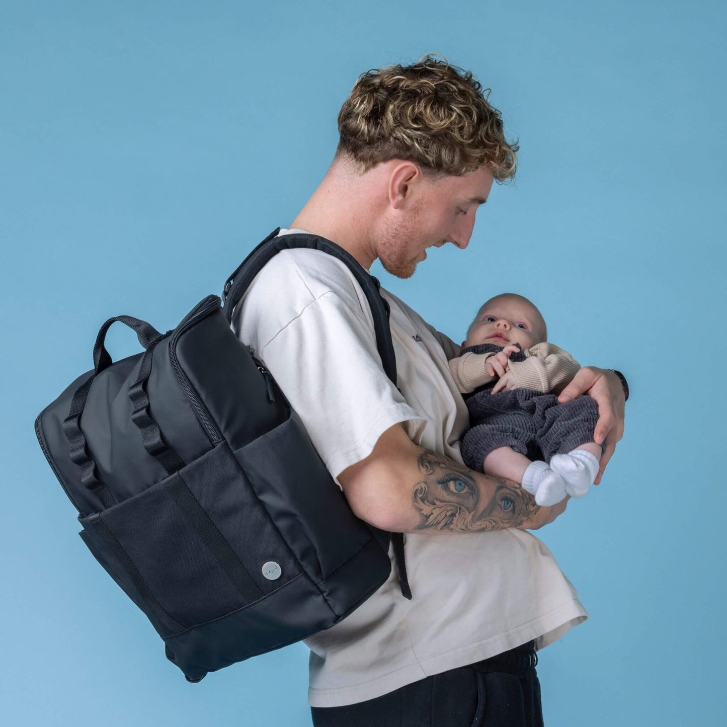 Parent wearing the Babymel Finn Changing Backpack in Black while smiling and holding a baby, showcasing the backpack's functional and stylish design for everyday parenting needs.