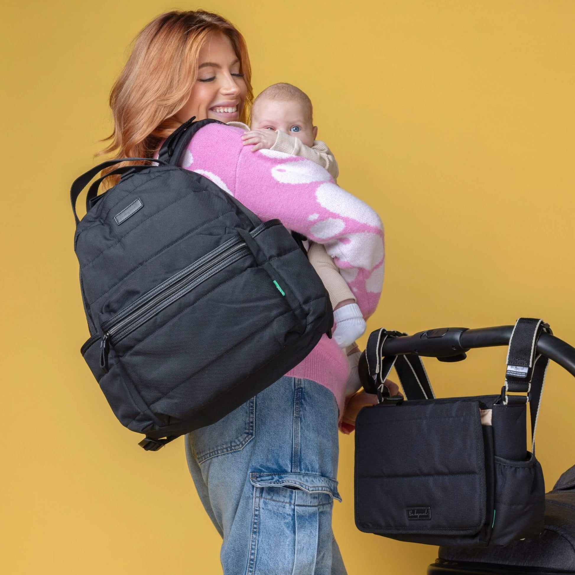 Parent carrying the Babymel Lola Eco Backpack in Black while holding a baby, with a matching stroller bag attached, showcasing versatility.