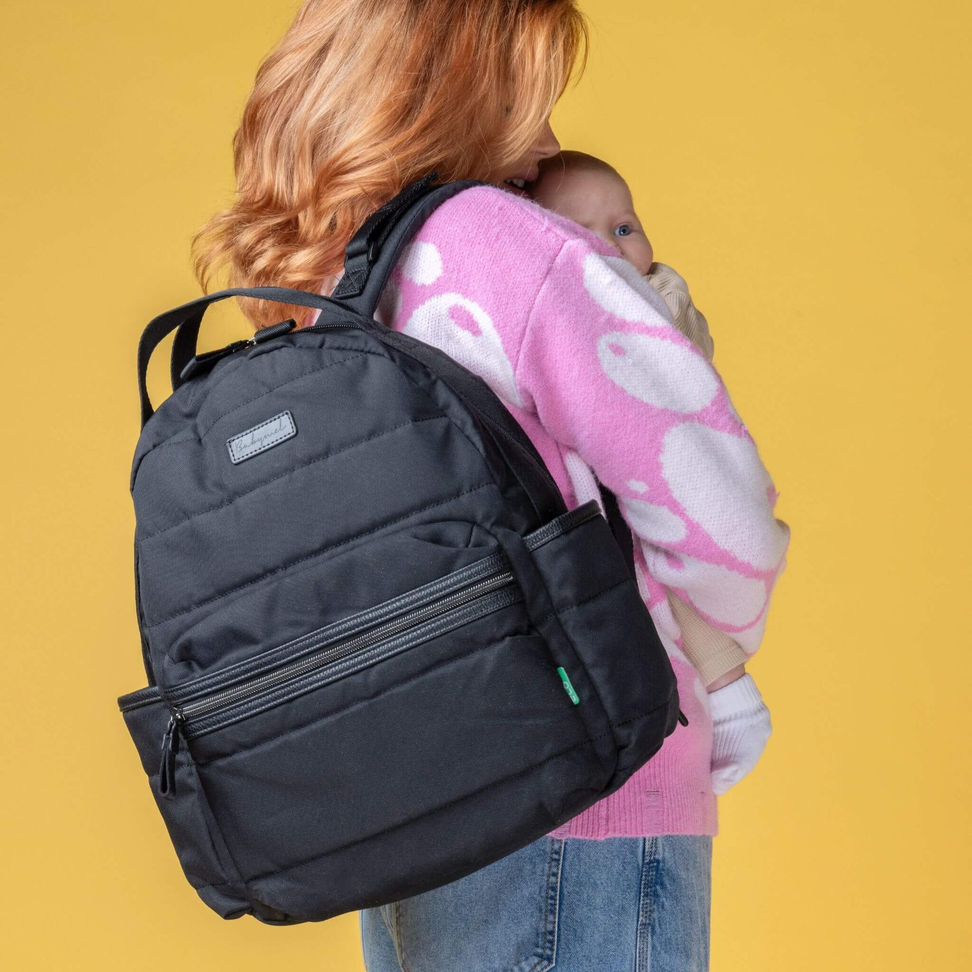 Mother carrying the Babymel Lola Eco Backpack in Black while holding a baby, showcasing its stylish and practical design for everyday use.