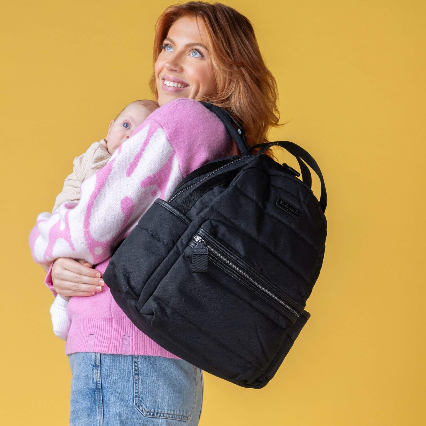 Smiling mother holding a baby while carrying the Babymel Lola Eco Backpack in Black, highlighting its comfortable and stylish design.