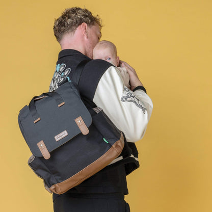 Father carrying baby while wearing the black Babymel Robyn Eco Convertible Backpack with tan details, showing its stylish and practical design.