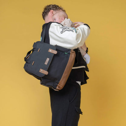 Father carrying baby while wearing the black Babymel Robyn Eco Convertible Backpack with tan details, styled as a shoulder bag for versatility.