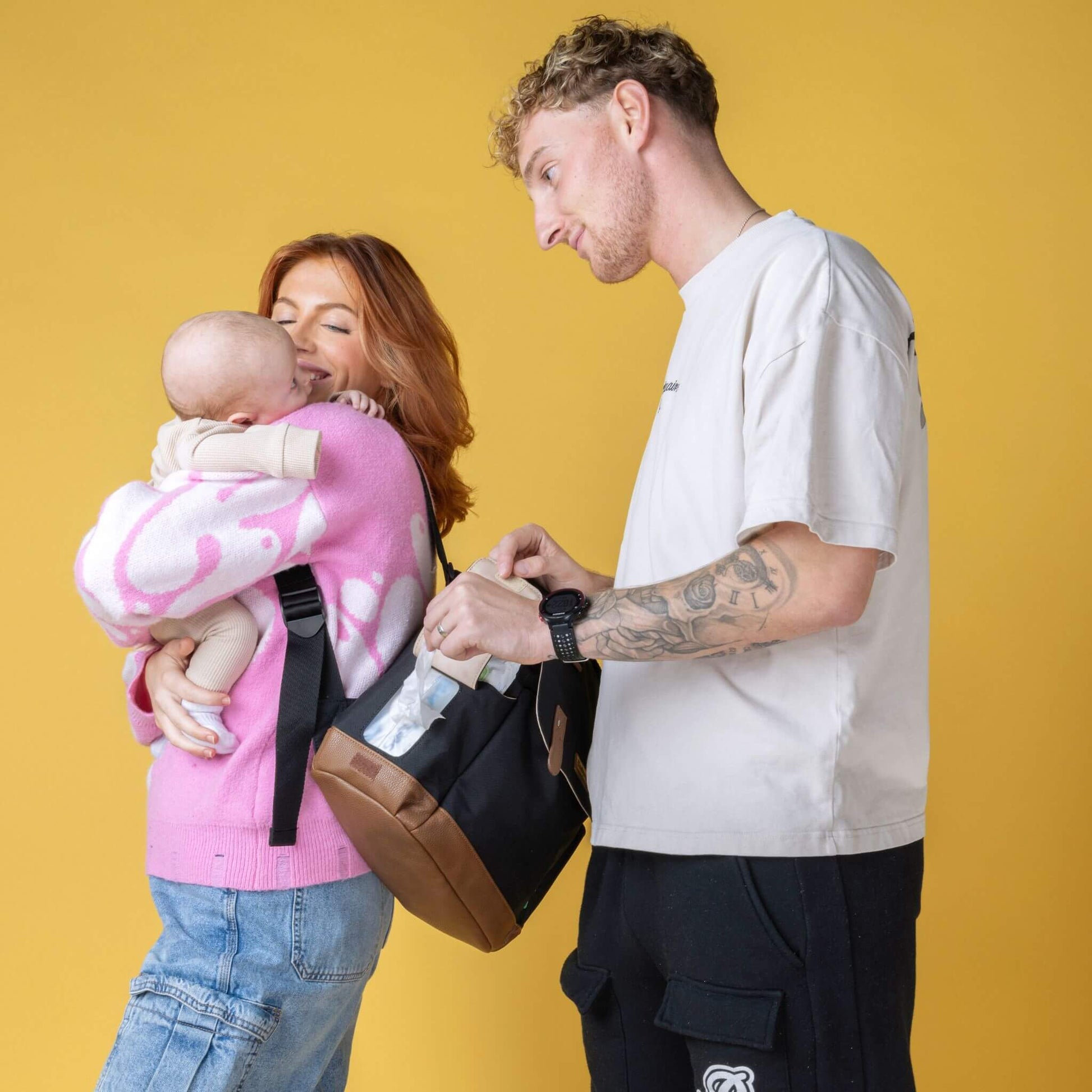 Parents with baby using the black Babymel Robyn Eco Convertible Backpack with tan details, highlighting the easy-wipes pocket for quick clean-ups.