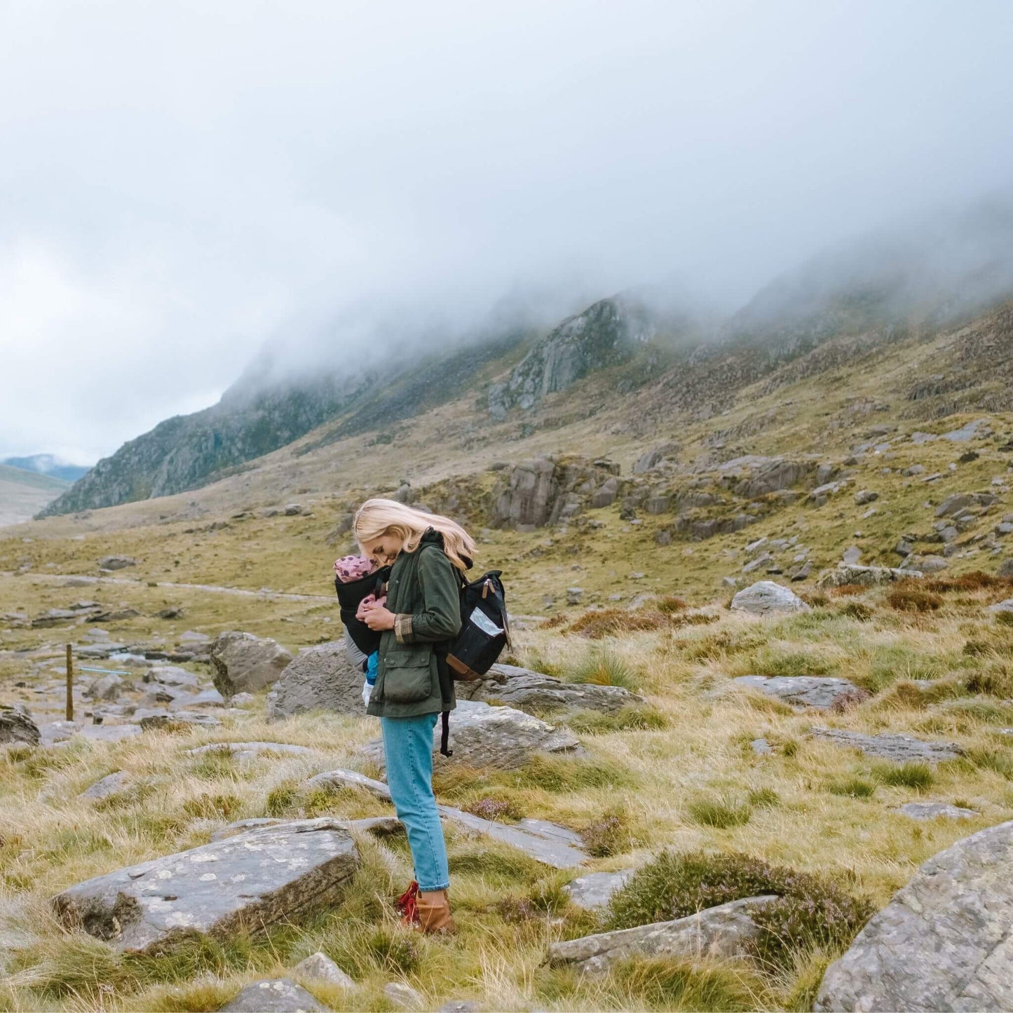 Mother carrying baby in a scenic outdoor setting, wearing the black Babymel Robyn Eco Convertible Backpack with tan details, highlighting its practicality for adventures.