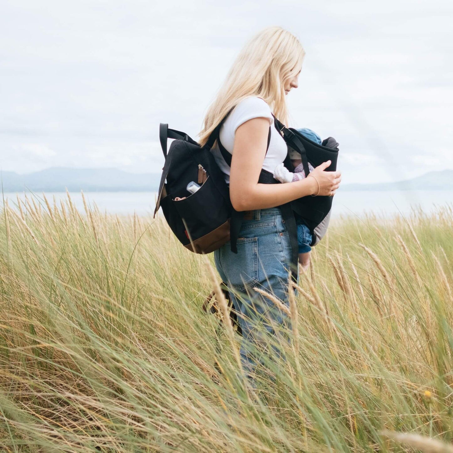 Mother carrying baby in a grassy coastal setting, wearing the black Babymel Robyn Eco Convertible Backpack with tan details, highlighting its practicality for outdoor adventures.