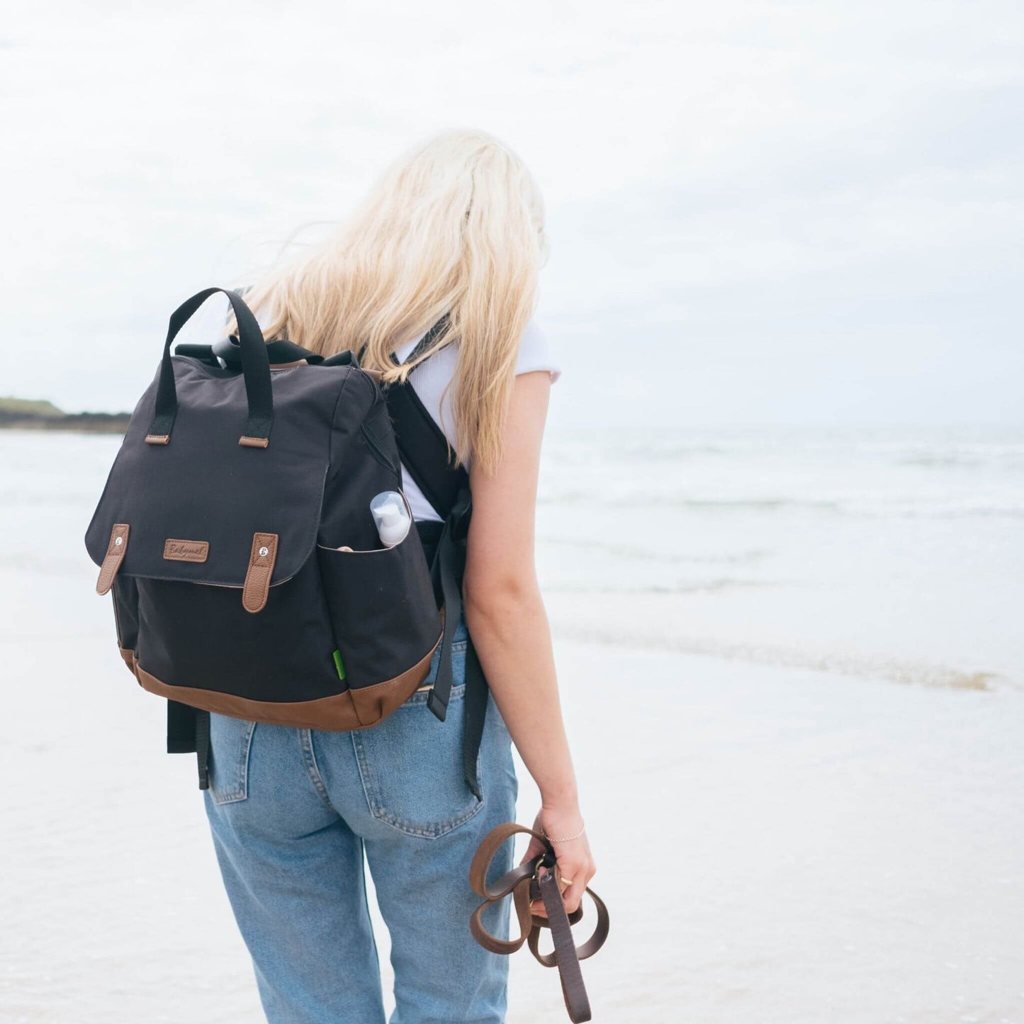 Mother walking along the beach wearing the black Babymel Robyn Eco Convertible Backpack with tan details, showcasing its stylish and practical design for travel.