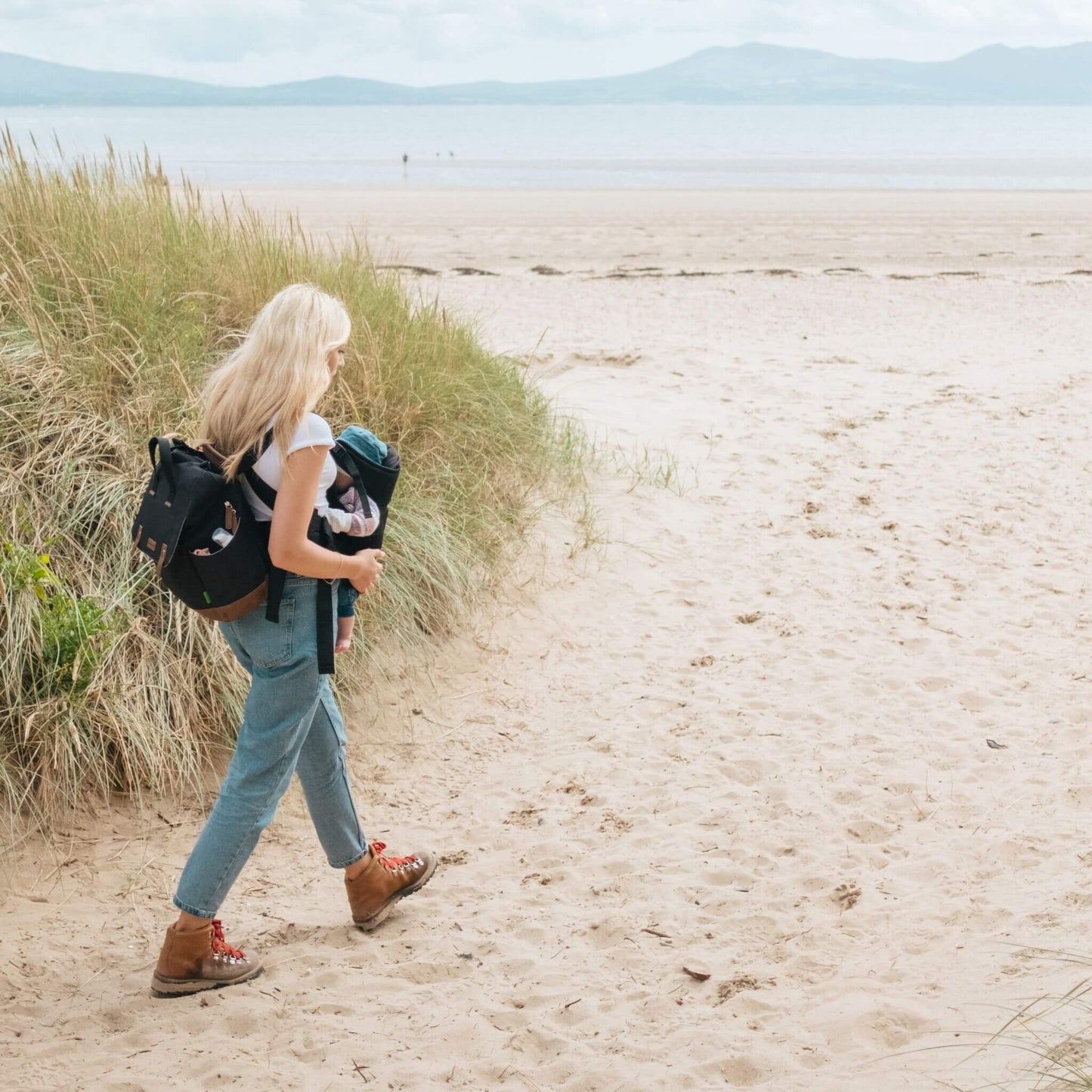 Mother carrying baby while walking on a sandy beach, wearing the black Babymel Robyn Eco Convertible Backpack with tan details, highlighting its practicality for outdoor adventures.