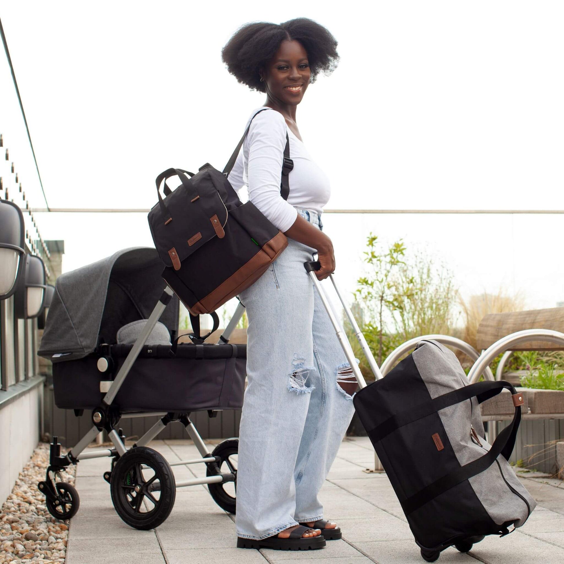 Smiling mother wearing the black Babymel Robyn Eco Convertible Backpack with tan details as a backpack, pulling a travel bag with a pram nearby, highlighting its stylish and practical design for travel.