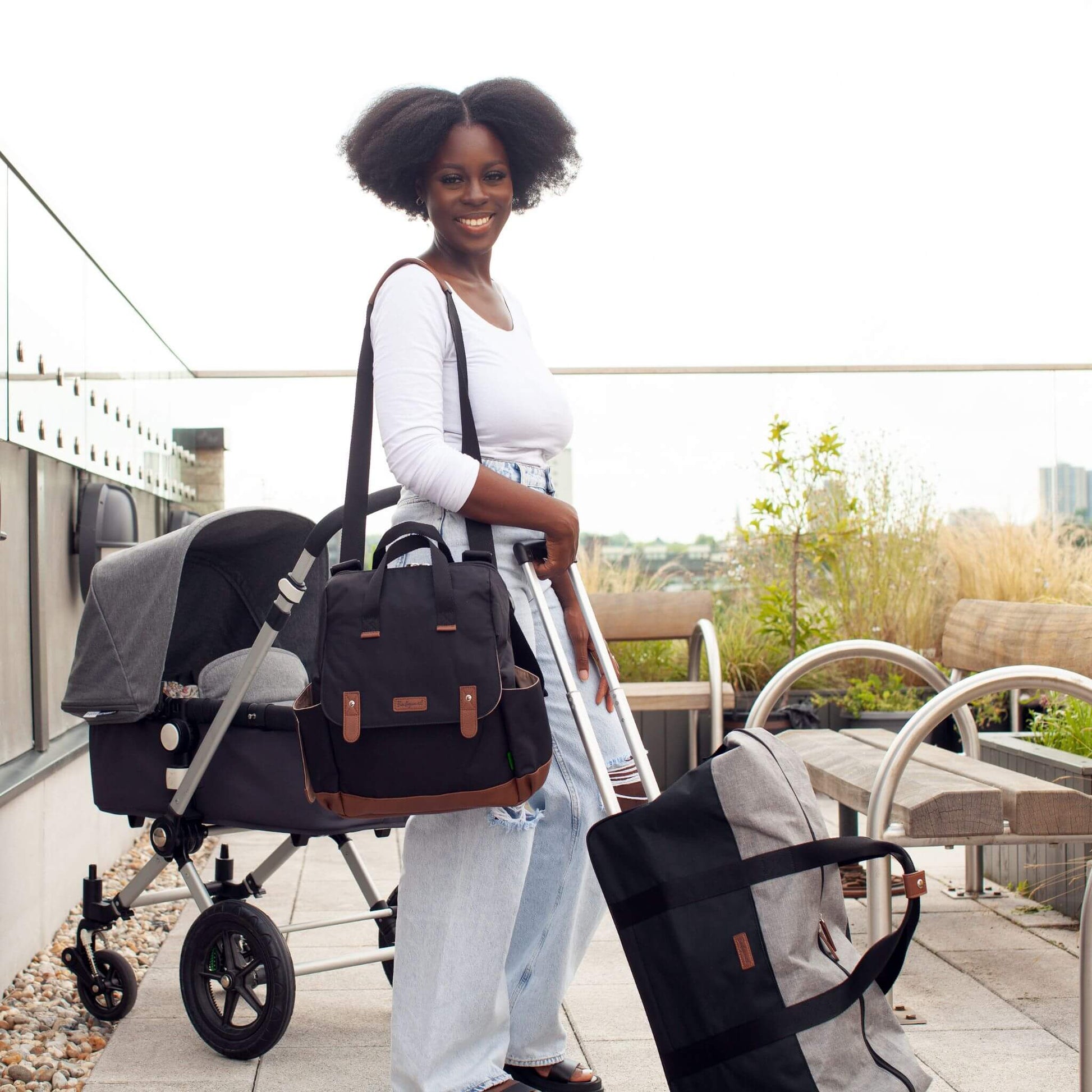 Smiling mother carrying the black Babymel Robyn Eco Convertible Backpack with tan details as a shoulder bag, with a pram and travel bag nearby, highlighting its stylish and versatile design for parents on the go.