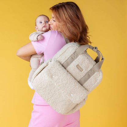 Mother holding baby while wearing the cream Babymel Teddy Convertible Backpack with soft borg fleece, highlighting its cosy and practical design.