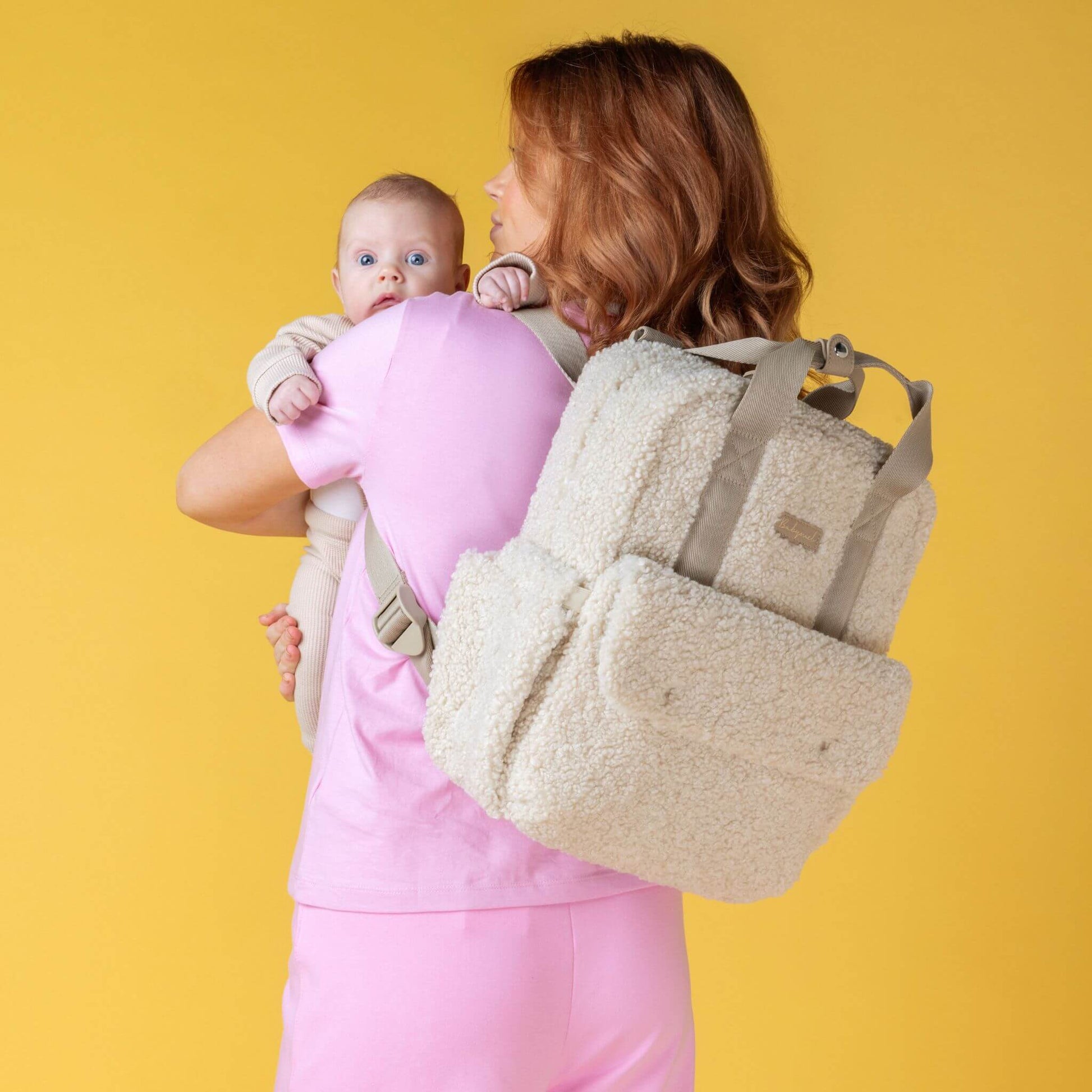 Mother holding baby while wearing the cream Babymel Teddy Convertible Backpack with soft borg fleece, styled as a backpack, showcasing its cosy and functional design.