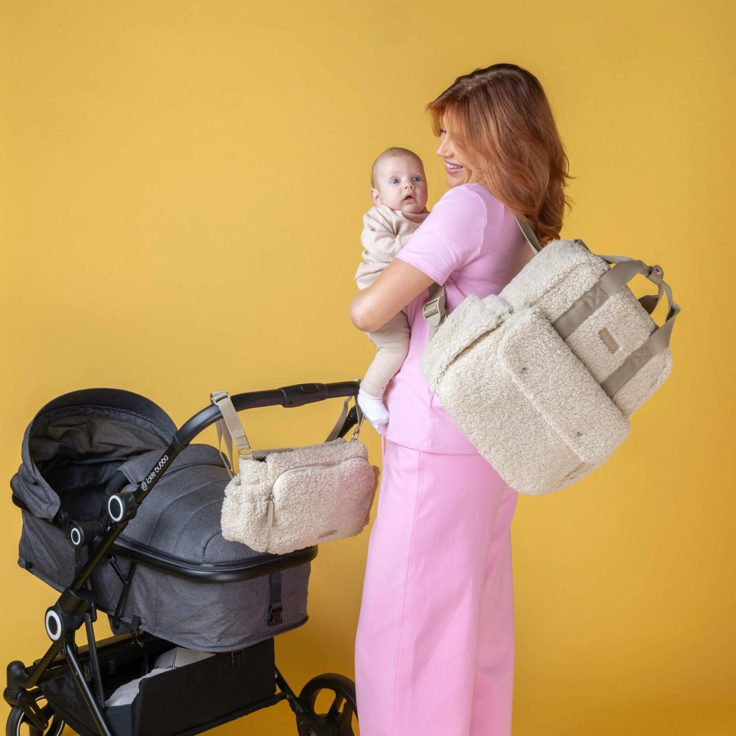 Smiling mother holding baby while wearing the cream Babymel Teddy Convertible Backpack with soft borg fleece, with a matching stroller organiser attached to a pram, showcasing the coordinated set.