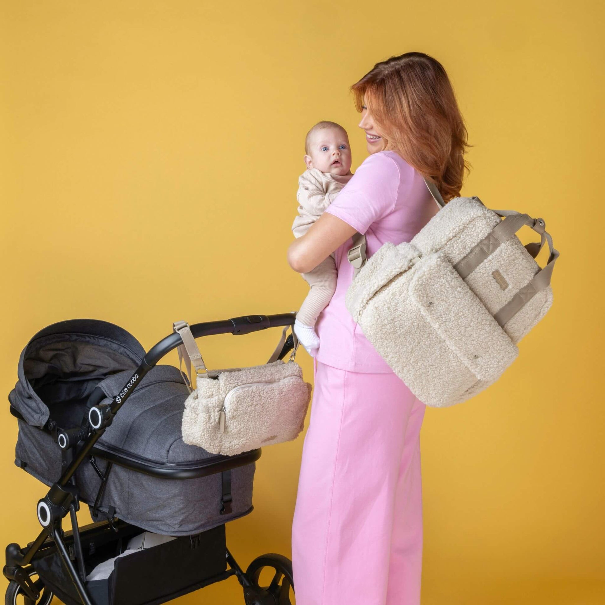 Smiling mother holding baby while wearing the cream Babymel Teddy Convertible Backpack with soft borg fleece, with a matching stroller organiser attached to a pram, showcasing the coordinated set.