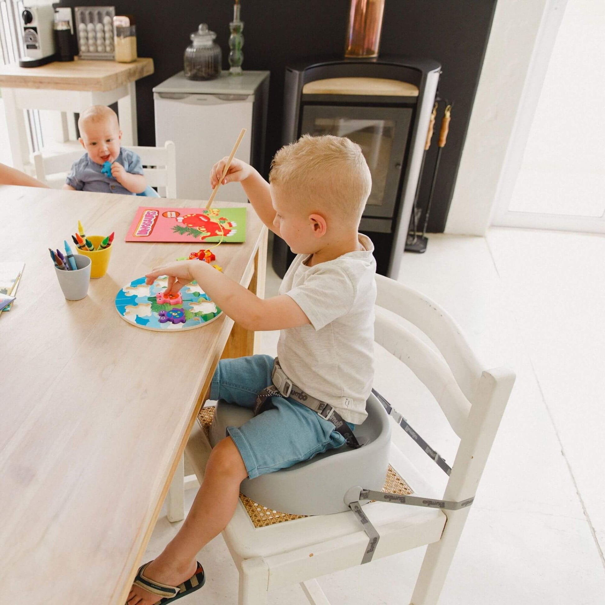 Young child seated in a Bumbo booster seat in cool grey, painting at a table with art supplies, securely strapped to the chair.