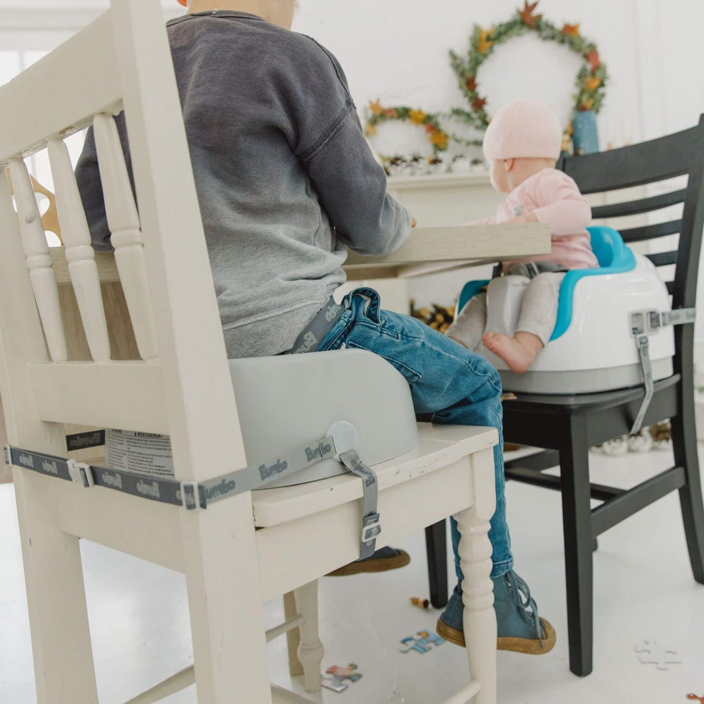 Two children seated at a table; older child in a cool grey Bumbo booster seat, younger child in a Bumbo seat, both securely strapped.