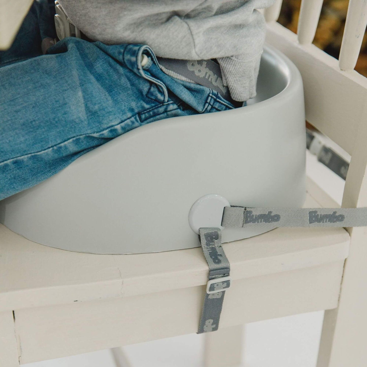 Close-up of a child seated in a cool grey Bumbo booster seat, securely strapped to a chair for stability and safety.