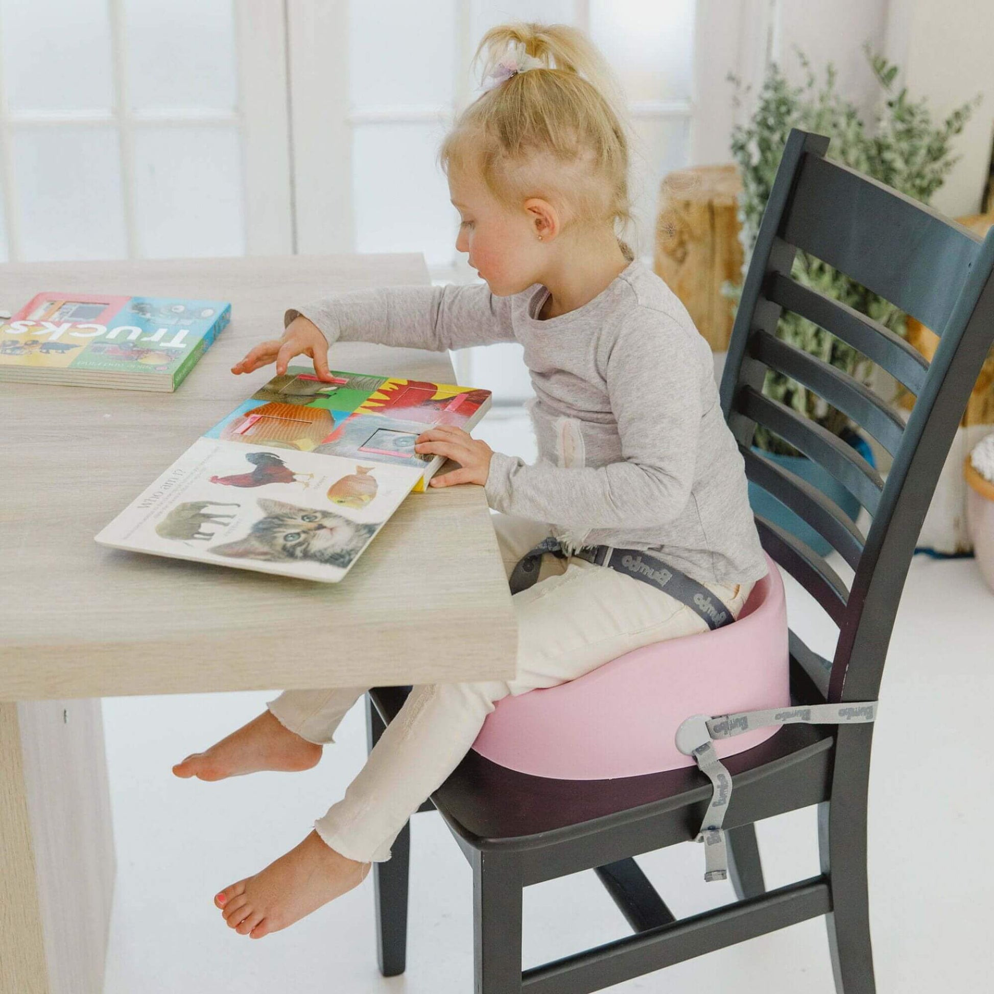 Young girl reading a book at a table while seated in a cradle pink Bumbo booster seat, securely strapped to a chair for safety.