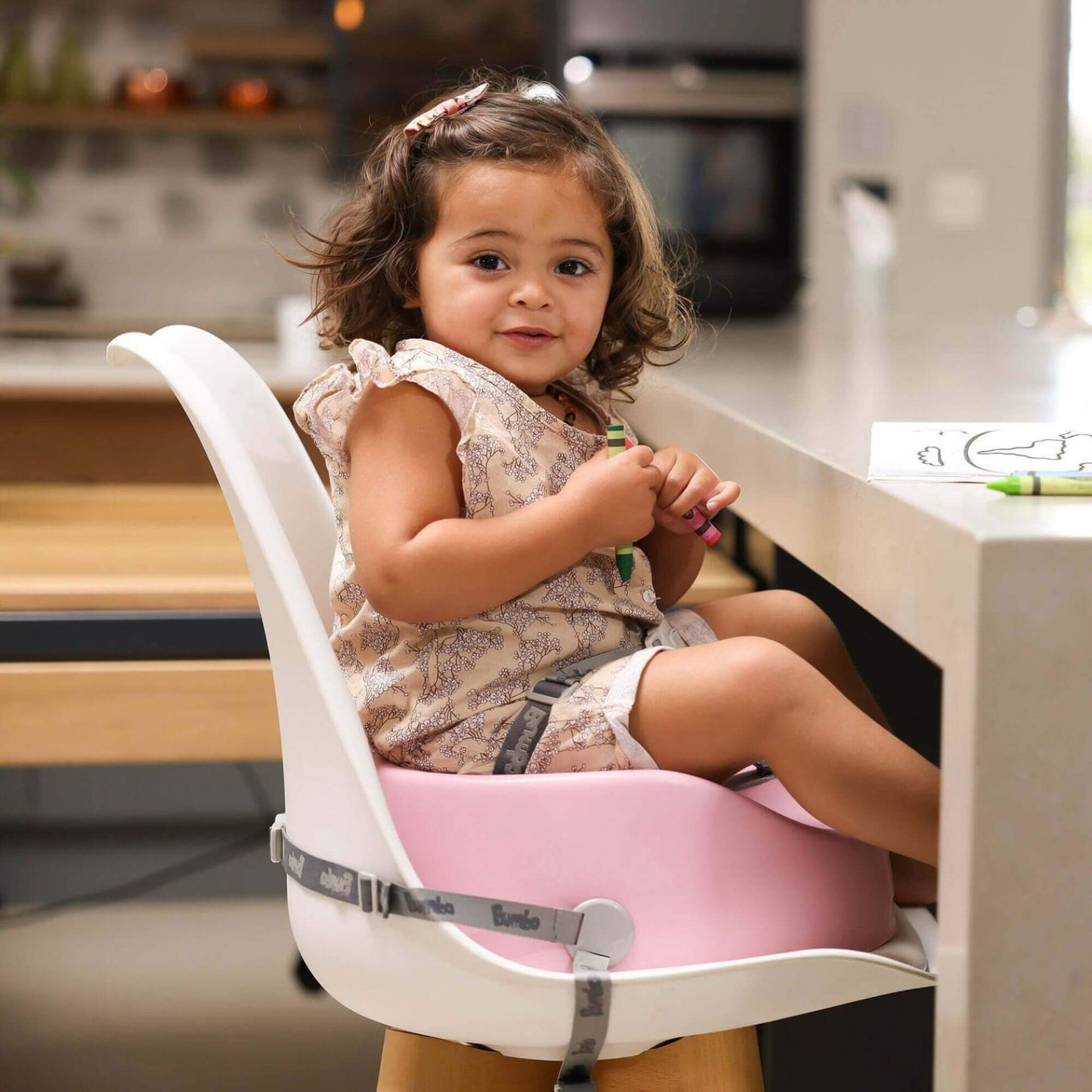 Toddler seated in a pink Bumbo booster seat, strapped securely to a chair, holding crayons and colouring at a table.