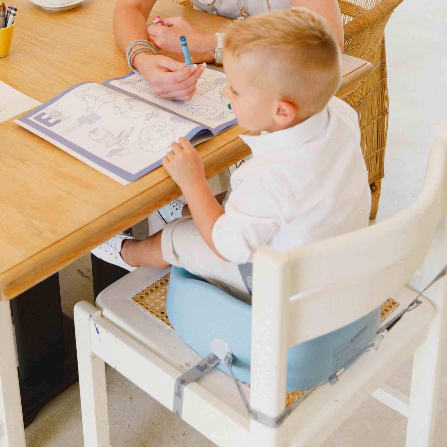 Toddler seated in a powder blue Bumbo booster seat, strapped securely to a chair, and colouring at a table.