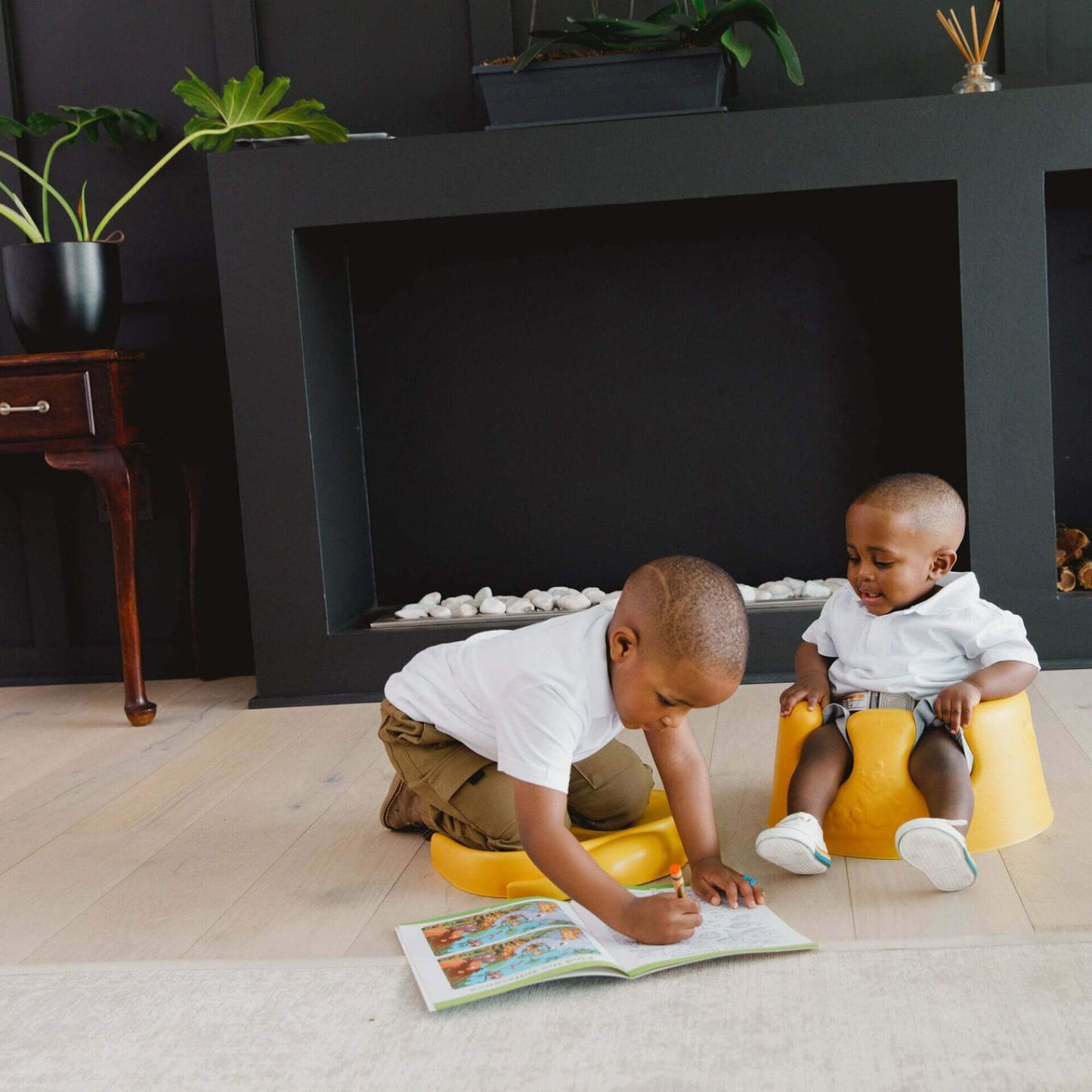 Two young children indoors; one kneeling on a mimosa yellow Bumbo Elipad, drawing in a book, while the other sits in a mimosa yellow Bumbo floor seat, watching.