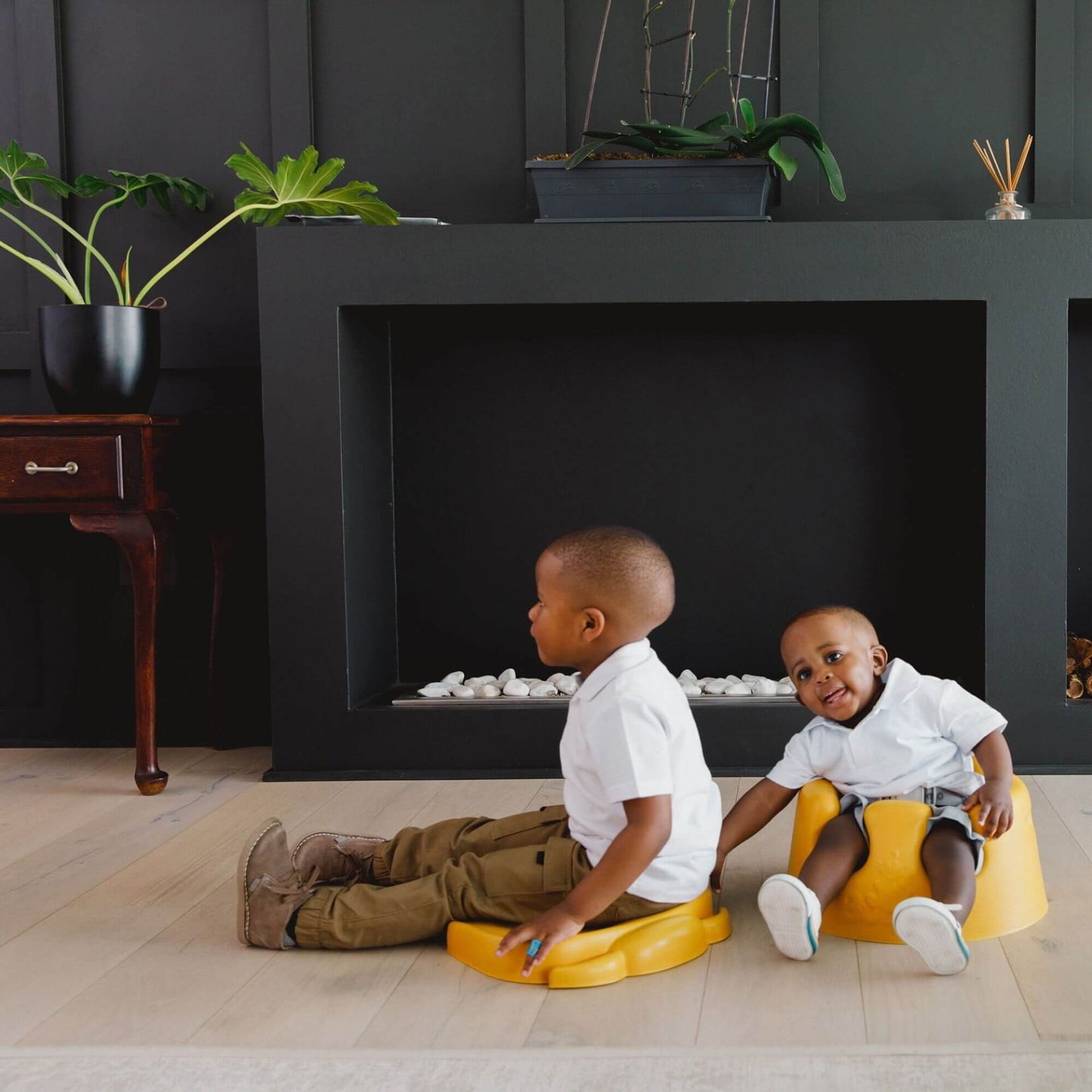 Two young children playing indoors, one sitting on a mimosa yellow Bumbo Elipad and the other in a mimosa yellow Bumbo floor seat, both smiling.