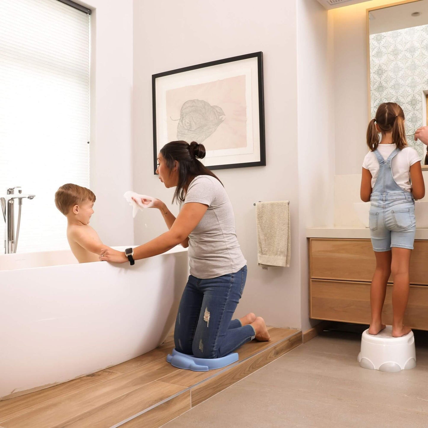 Mother kneeling on a powder blue Bumbo Elipad while bathing her child, with another child using a step stool to reach the bathroom sink.