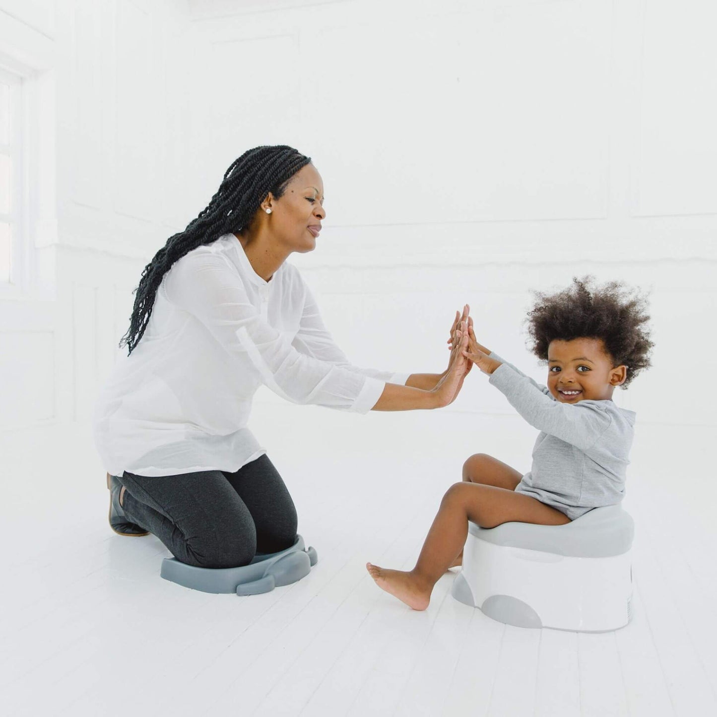 Mother kneeling on a slate grey Bumbo Elipad, giving a high-five to her smiling toddler seated on a training potty.