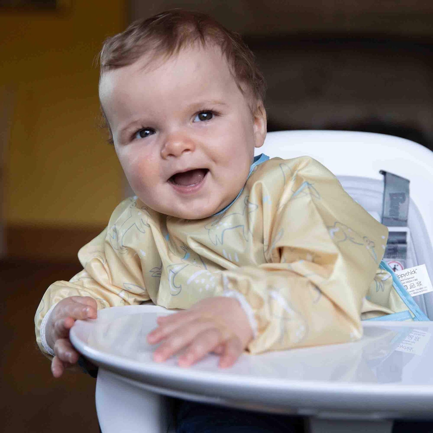 Smiling baby in a Bumbo highchair wearing a yellow bib, comfortably seated with hands on the detachable tray, ready for mealtime.