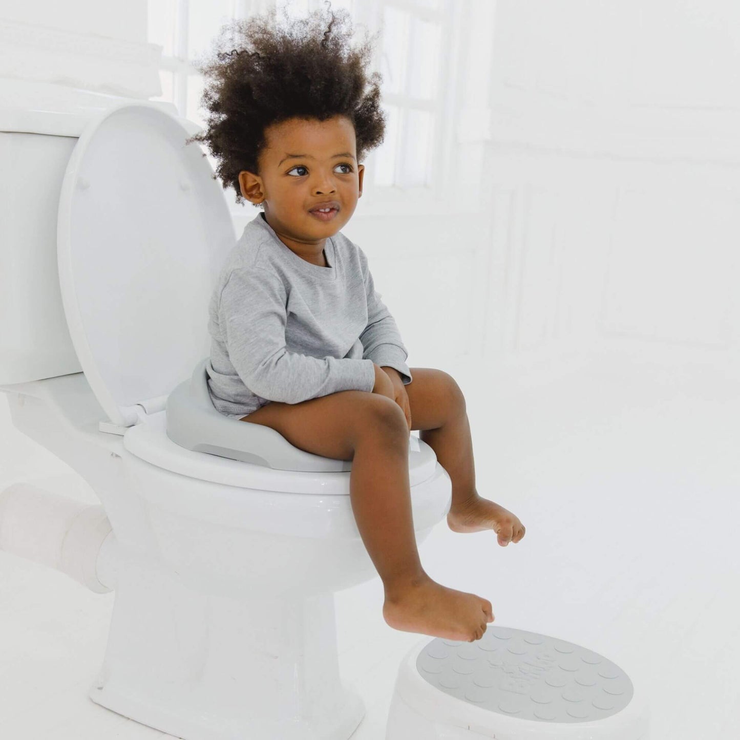 Toddler sits on a cool grey Bumbo toilet trainer, with a matching step stool nearby, making toilet training comfortable and accessible.