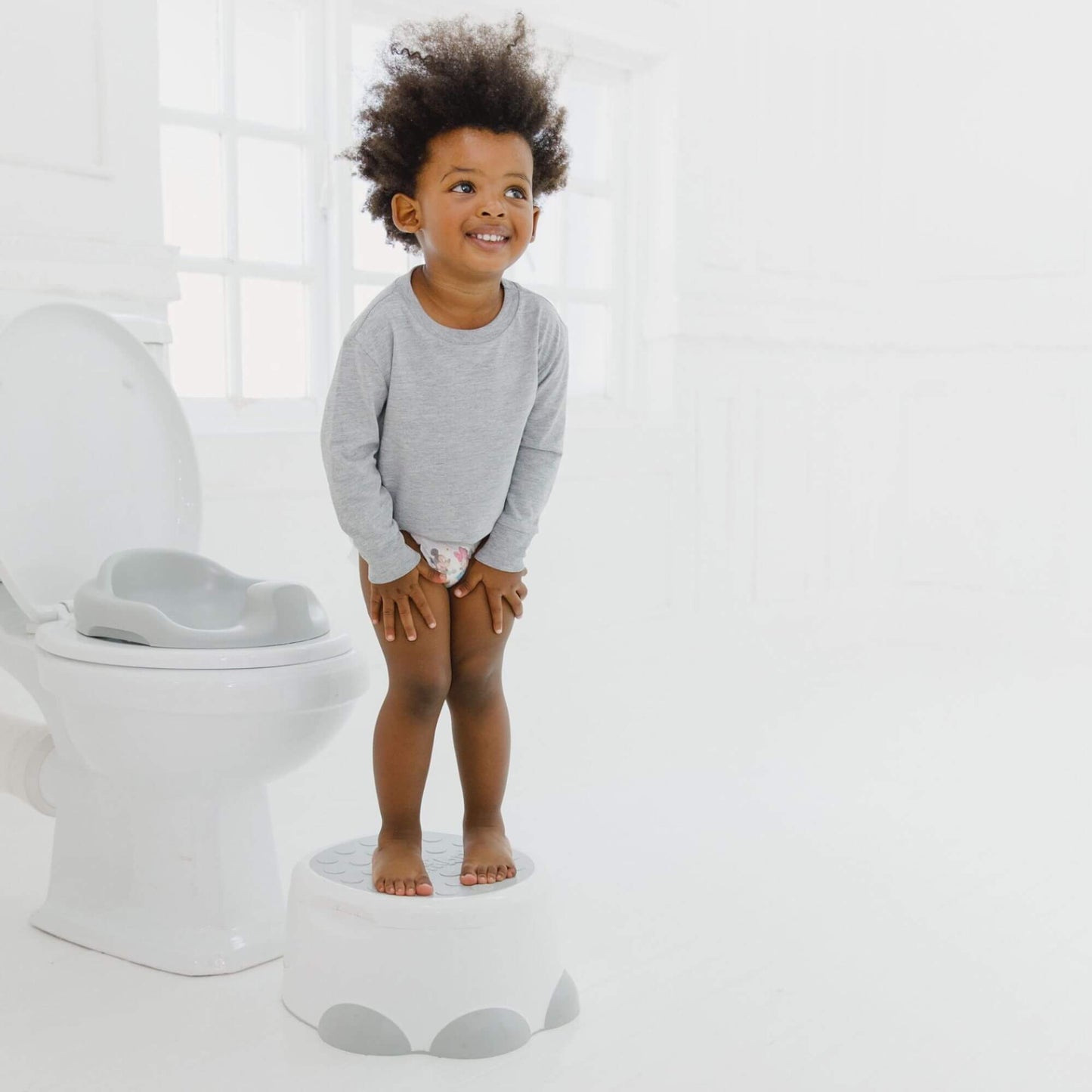 Smiling toddler stands on a cool grey Bumbo step stool next to a toilet with a Bumbo toilet trainer seat, ready for independent bathroom use.