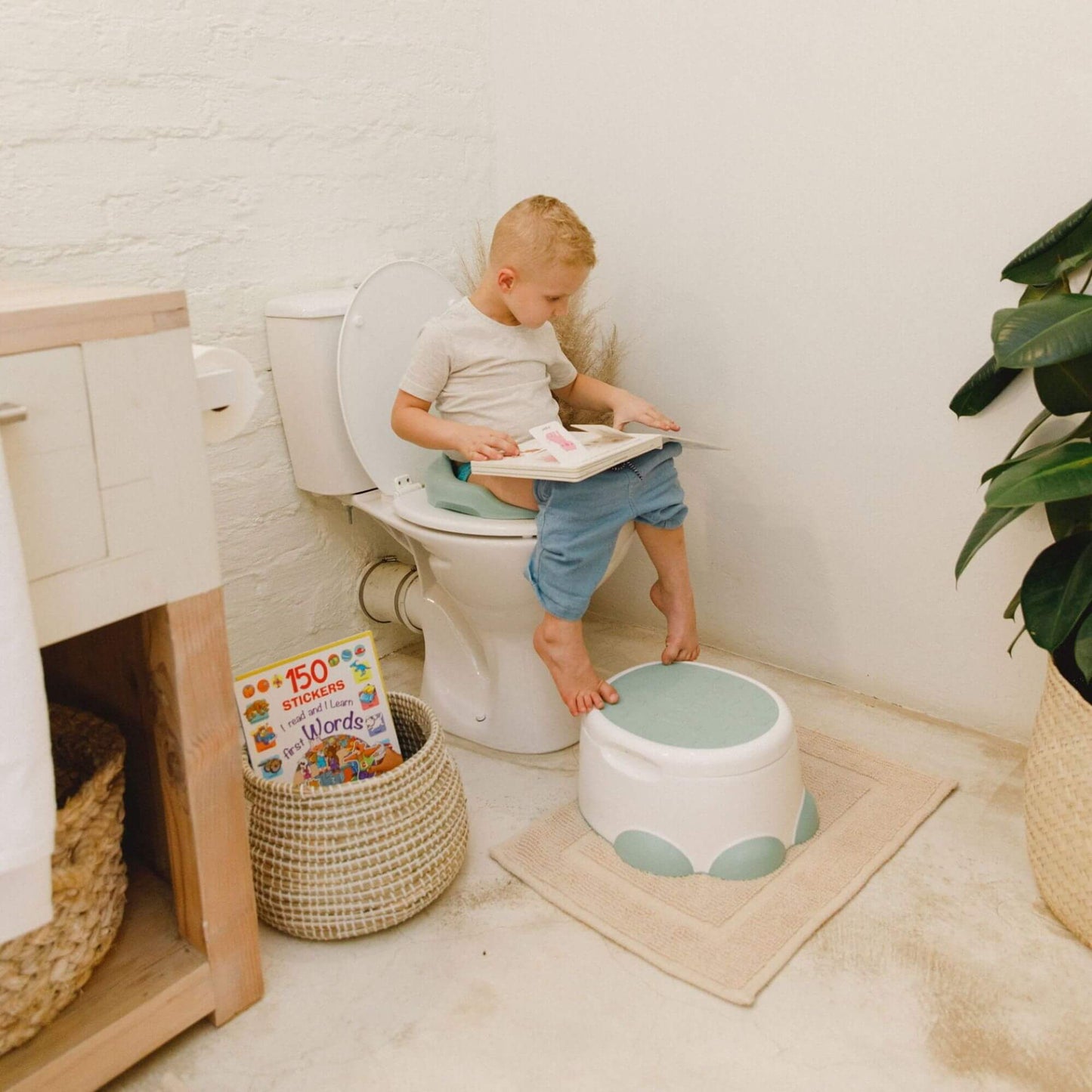 Child sits on a Bumbo hemlock green toilet trainer, reading a book on the toilet with a matching step stool nearby.