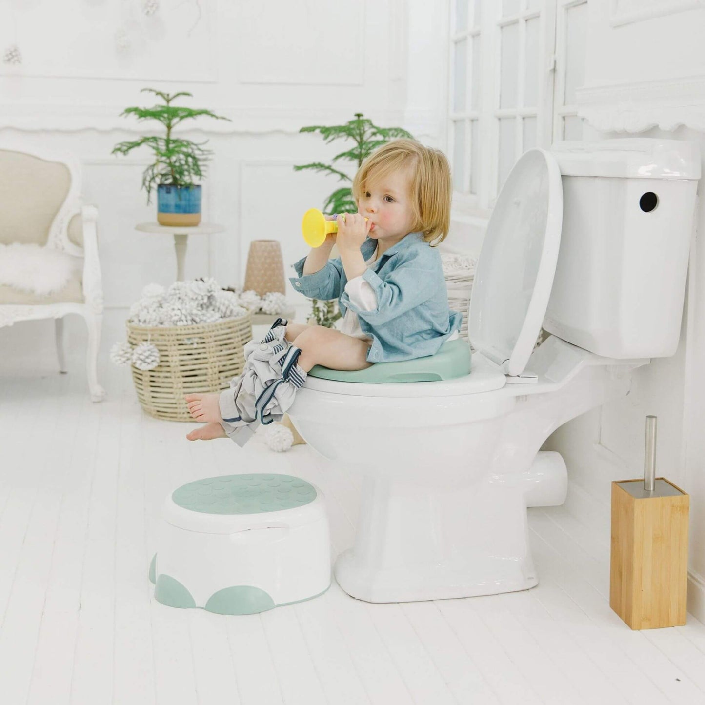 Toddler sits on a Bumbo toilet trainer in light green on a toilet, playing with a yellow trumpet, with a step stool nearby.