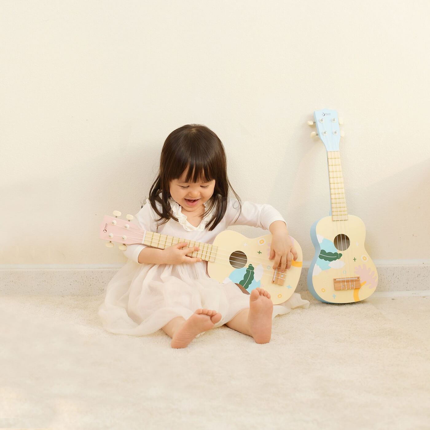 A young girl happily playing the pink Classic World Iris Ukulele, with a matching blue ukulele resting nearby, inspiring creative fun.