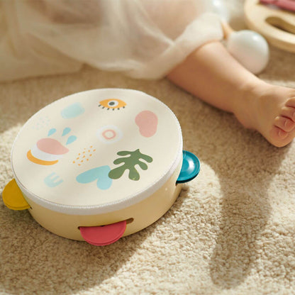 A tambourine from the Classic World Iris Music Set, featuring playful designs, resting on soft carpet near a child’s foot.