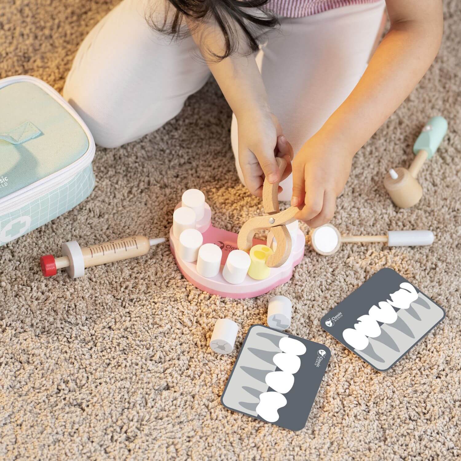 A child uses the Classic World Little Dentist Set, holding wooden pliers to remove a pretend tooth from a dental model on a carpeted floor.