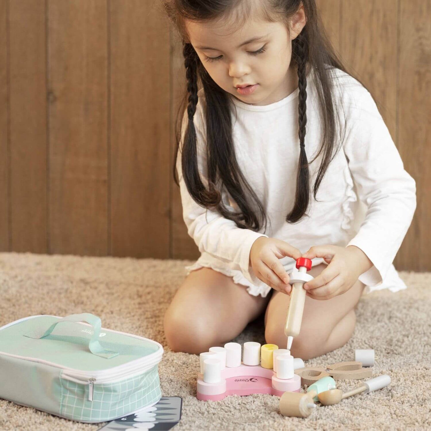 A child sits on a carpeted floor, using the Classic World Little Dentist Set, holding a wooden syringe with the tools and dental set nearby.