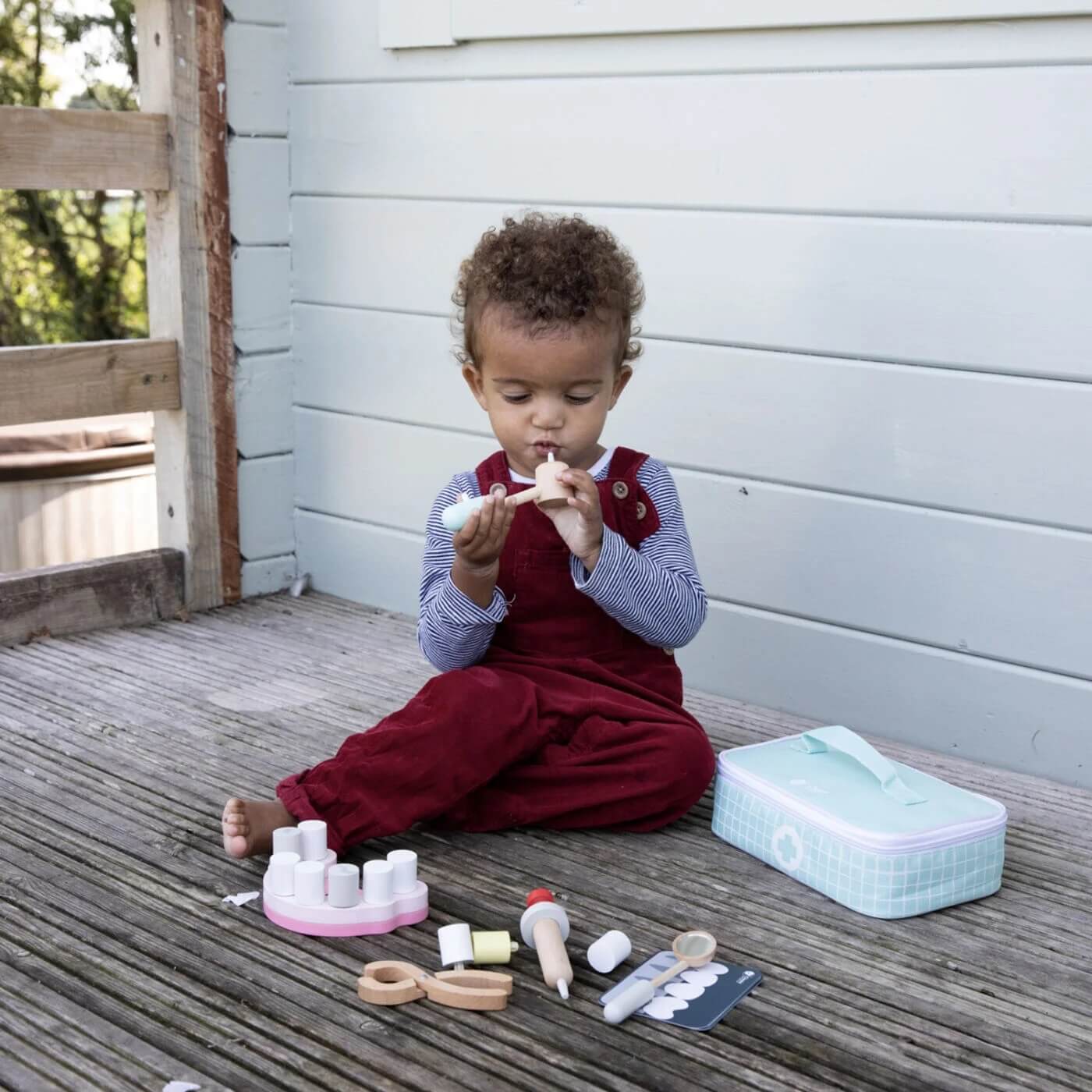 A child sits on a wooden deck, focused on playing with the Classic World Little Dentist Set, holding a dental tool with the kit spread around.