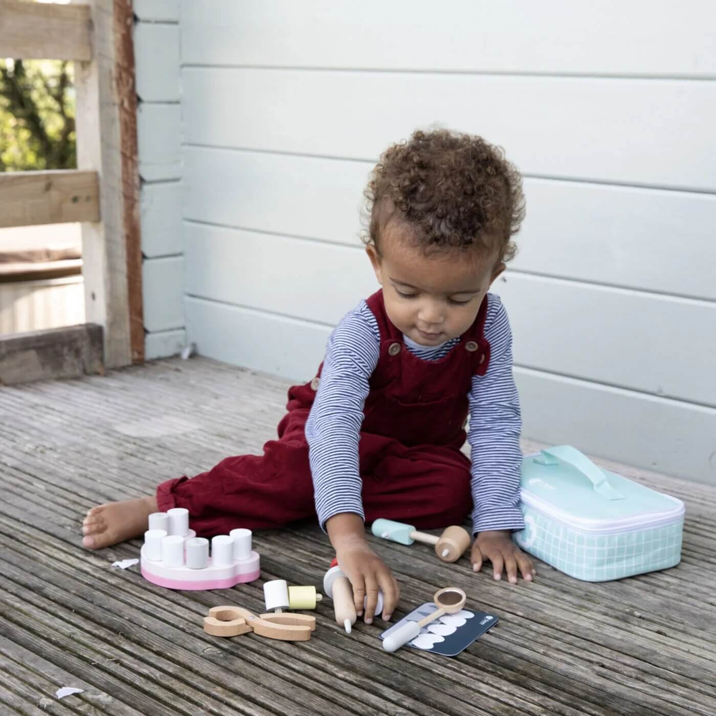 A child sits on a wooden deck, exploring the Classic World Little Dentist Set, surrounded by wooden dental tools, teeth, and a carry case.