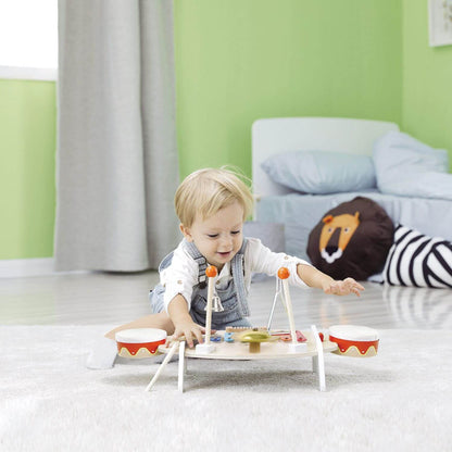 A toddler joyfully plays on the floor with the Classic World Music Table, exploring its instruments in a bright, cosy room.