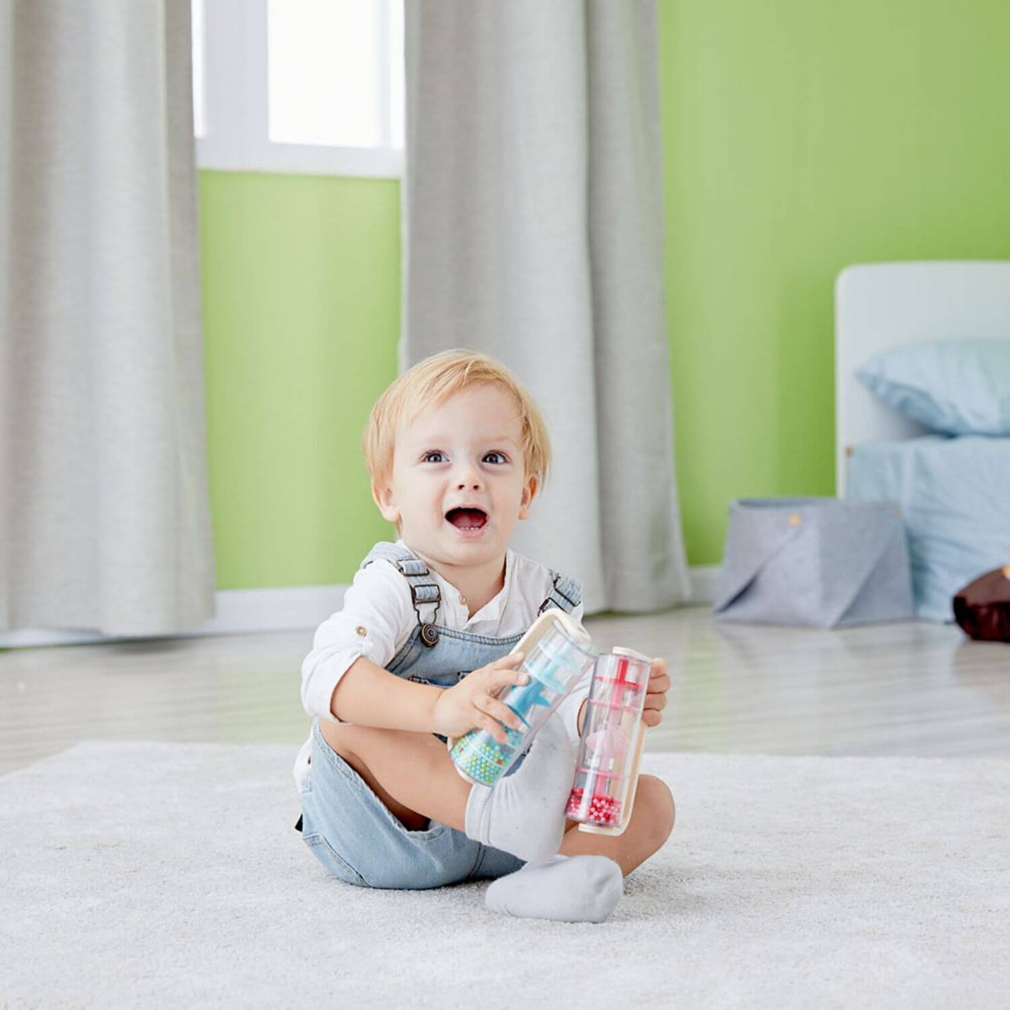 A smiling toddler sitting on a soft carpet, playing with the Classic World Rainmaker toys, featuring colourful beads and wooden handles.