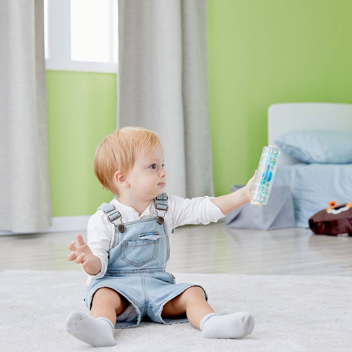 A toddler sitting on a carpet, holding up the Classic World Rainmaker toy, watching the colourful beads cascade inside with curiosity.