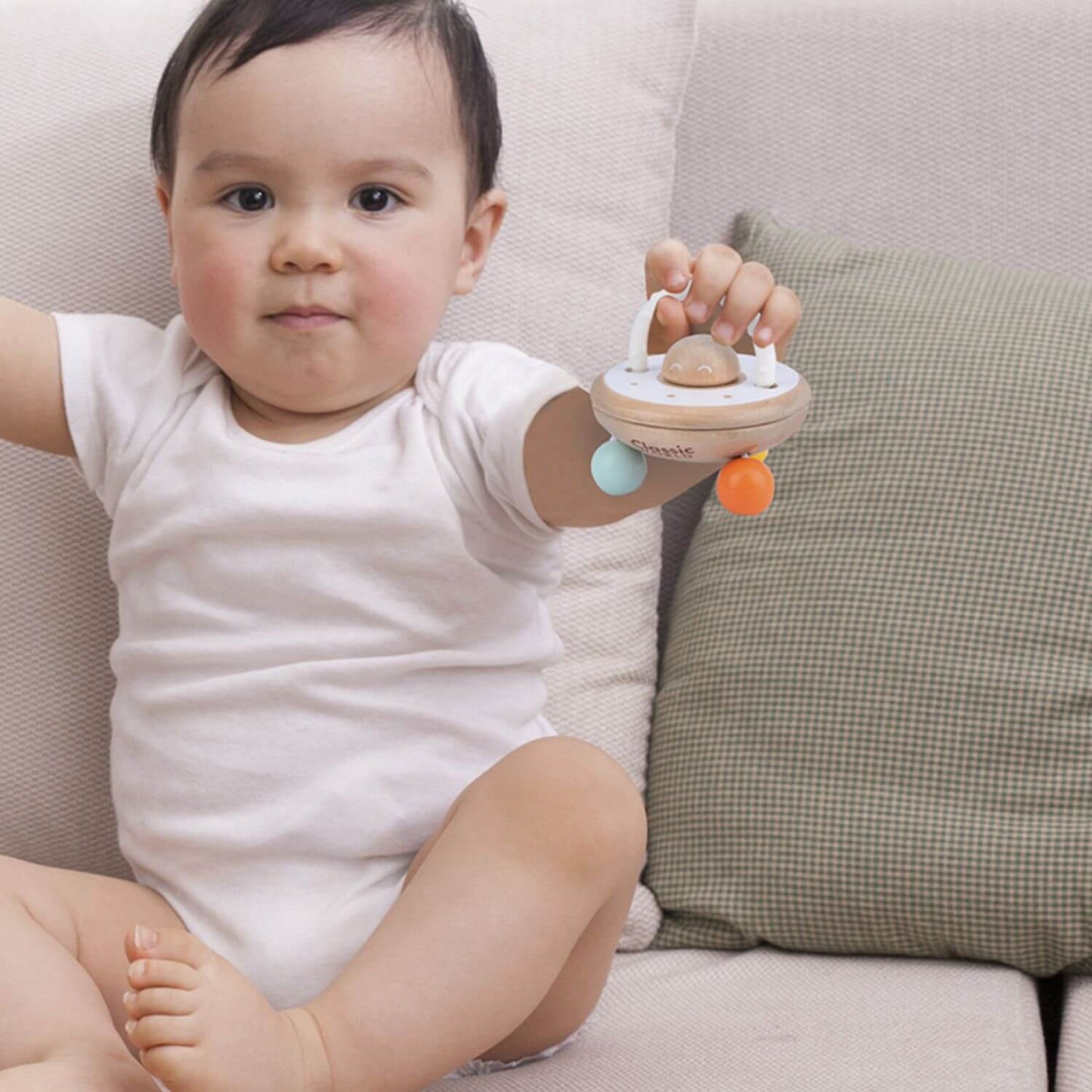 A baby sitting on a sofa holding the Classic World UFO Rattle, showcasing its easy-to-grasp design and engaging features for little hands.