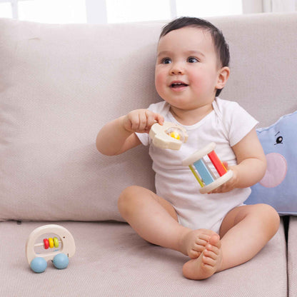 Smiling baby playing with Classic World roller rattle, sitting on a sofa, surrounded by soft toys, encouraging sensory exploration.