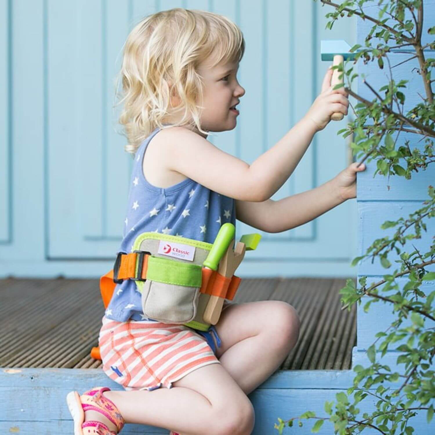 A young girl wearing the Classic World Tool Belt with wooden tools, playfully hammering outdoors, showcasing its creative and portable design.