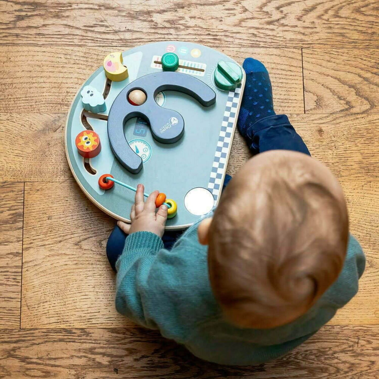 Toddler exploring the Classic World Travel Busy Board on the floor, engaging with features like the steering wheel and bead slider.