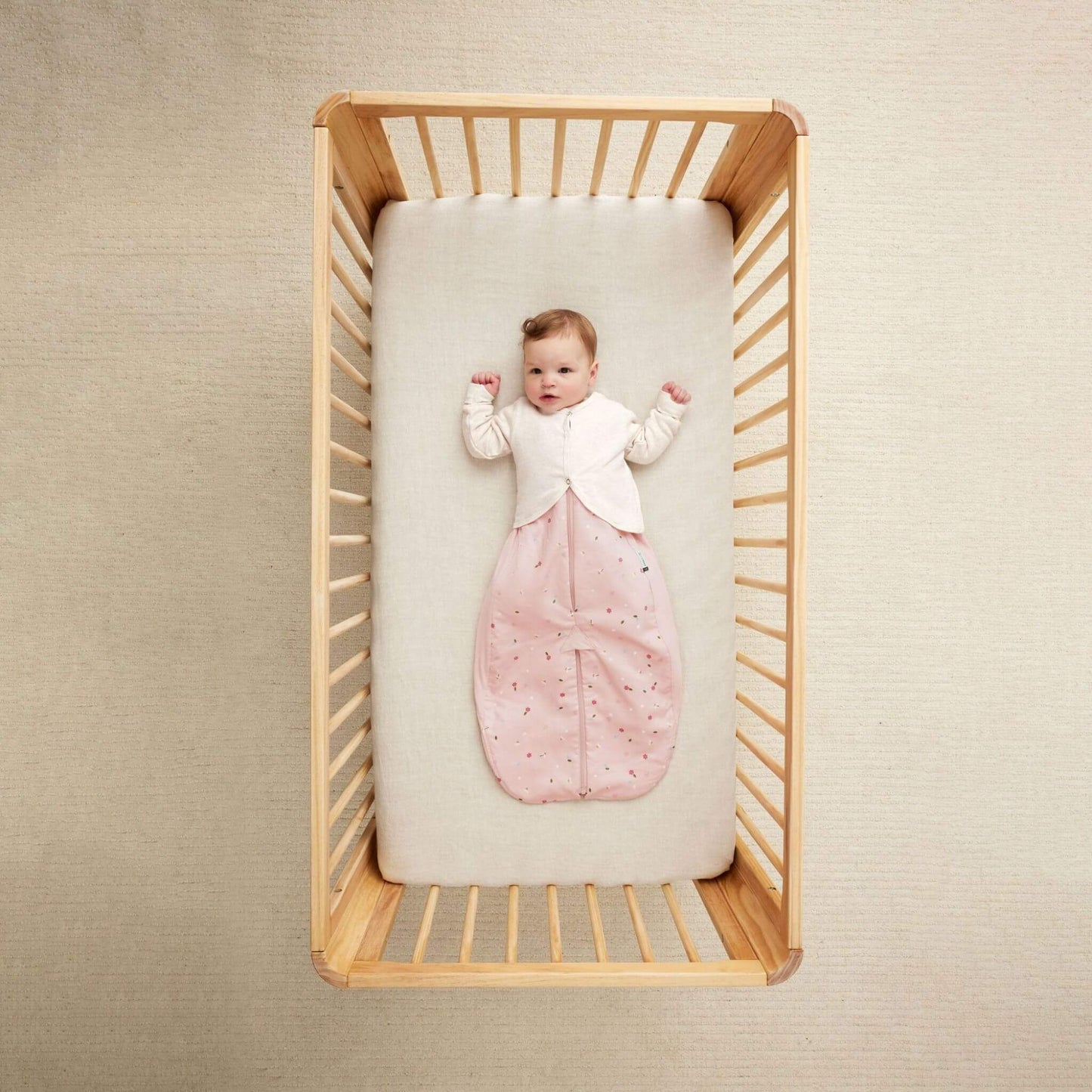 Baby lying in a cot wearing the ErgoPouch Long Sleeved Arm Warmer in oatmeal over a pink sleeping bag. A cosy, breathable layer for added warmth.