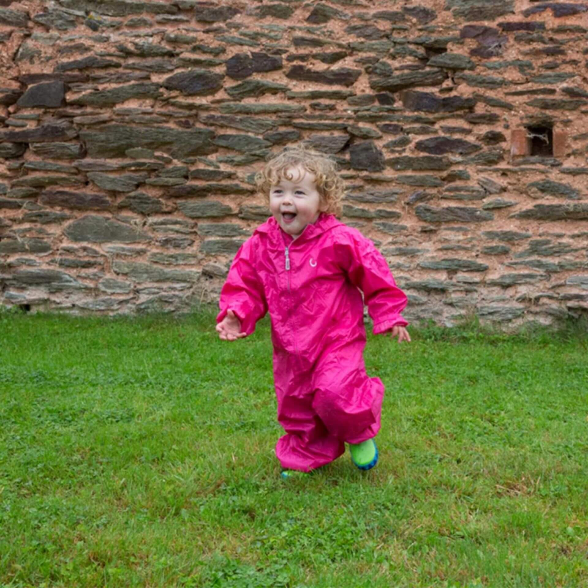 A joyful toddler in a pink rain suit and blue/green wellington boots runs on grass in front of an old stone wall, smiling widely.